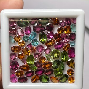 May include: A collection of colorful gemstones in a white display case. The gems are various shapes and sizes, including oval, pear, and round cuts. Colors include pink, green, orange, and blue. The gems are arranged closely together.