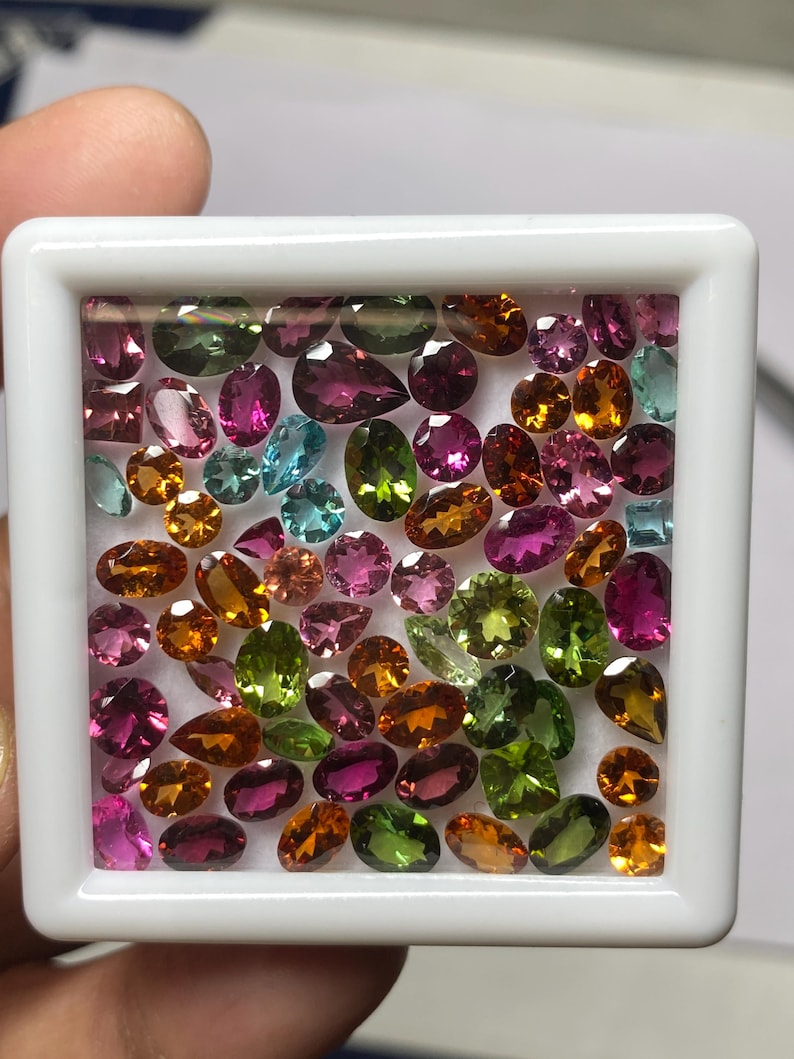 May include: A collection of colorful gemstones in a white display case. The gems are various shapes and sizes, including oval, pear, and round cuts. Colors include pink, green, orange, and blue. The gems are arranged closely together.