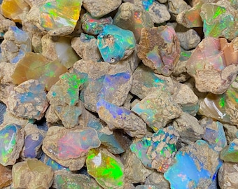 May include: A collection of raw opal stones, displaying a spectrum of colours such as blues, greens, yellows, and reds. The opals have a rough, natural texture and are set within a matrix of brown rock.