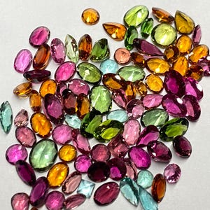 May include: A collection of colorful gemstones in various shapes and sizes. The gems are in shades of pink, green, orange, yellow, and blue. The gemstones are scattered on a white surface, creating a vibrant display of color and light.
