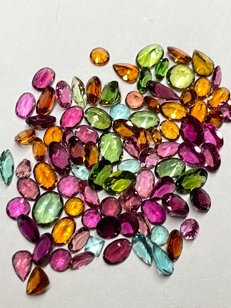 May include: A collection of colorful gemstones in various shapes and sizes. The gems are in shades of pink, green, orange, yellow, and blue. The gemstones are scattered on a white surface, creating a vibrant display of color and light.