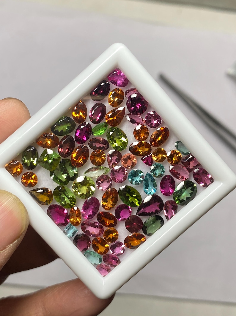 May include: A collection of colorful gemstones in various shapes and sizes, including oval, pear, and round cuts. The gems are displayed in a white square tray, showcasing a range of colors from green and blue to pink and orange.