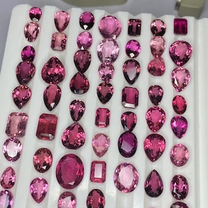 May include: A selection of pink gemstones in diverse shapes, such as round, oval, pear, and rectangular cuts. The gemstones are displayed on a white surface, highlighting their varied shades and sizes. A measuring tool is visible in the background.