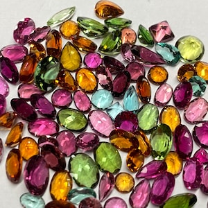 May include: A collection of colorful gemstones in various shapes and sizes. The gems are in shades of pink, green, yellow, orange, and blue. The gemstones are oval, teardrop, and round, with a polished, reflective surface.