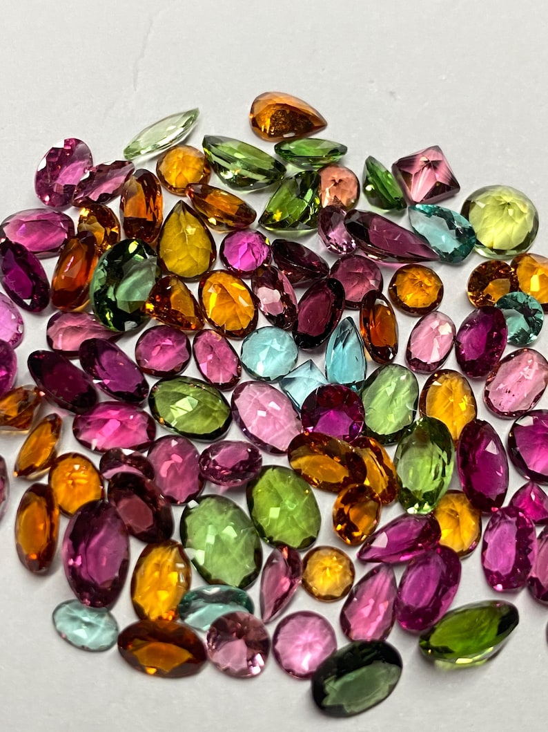 May include: A collection of colorful gemstones in various shapes and sizes. The gems are in shades of pink, green, yellow, orange, and blue. The gemstones are oval, teardrop, and round, with a polished, reflective surface.
