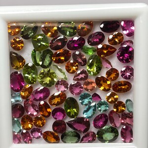 May include: A collection of colorful gemstones in a white tray. The gems are oval and teardrop-shaped, in shades of pink, green, orange, and blue. The gemstones are arranged closely together, filling the tray.