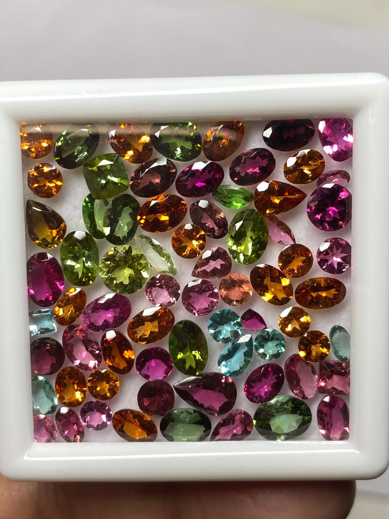 May include: A collection of colorful gemstones in a white tray. The gems are oval and teardrop-shaped, in shades of pink, green, orange, and blue. The gemstones are arranged closely together, filling the tray.