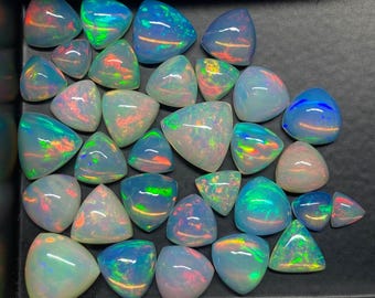 Puede incluir: Una colección de ópalos en varias formas, incluyendo triángulos y formas redondeadas. Los ópalos muestran un juego de color con destellos iridiscentes de azul, verde, naranja y rojo. Las piedras preciosas están sobre un fondo oscuro, resaltando sus vibrantes tonos.