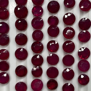 May include: A collection of round, faceted, deep red rubies arranged in neat vertical rows on a white grooved display tray. Each gemstone features a brilliant cut with multiple facets that reflect light, showcasing a consistent dark crimson hue.