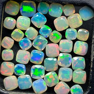 May include: A selection of square-cut opals, displaying a range of colours including green, blue, and pink. The gemstones are closely arranged, highlighting their polished surfaces and internal colour play. The opals vary in size and colour intensity.