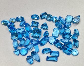 May include: A selection of bright blue gemstones in various shapes. The gems are scattered on a light grey surface, with cuts including oval, rectangular, and teardrop forms. The gemstones vary in size and reflect light, creating a sparkling effect.