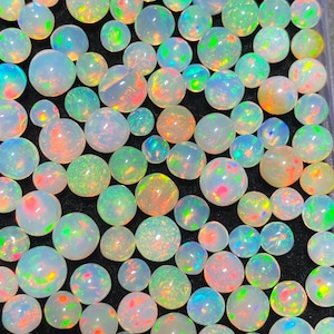 May include: A collection of opals in various sizes, arranged closely together. The opals display a range of colors, including white, blue, green, orange, and pink, with iridescent flashes. The gemstones are round and polished, set against a dark background.
