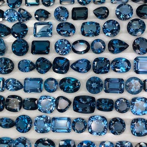 Könnte beinhalten: Eine Sammlung verschiedener blauer Edelsteine, darunter runde, ovale, quadratische und rechteckige Formen. Die Edelsteine sind auf einer weißen Oberfläche angeordnet, um ihre verschiedenen Schliffe und Facetten zu zeigen. Die Steine variieren in Blautönen.