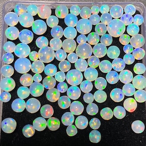 May include: A collection of round opals in various sizes, displayed in a clear plastic container. The opals exhibit a milky white base color with iridescent flashes of blue, green, and yellow. The opals are arranged on a black surface.