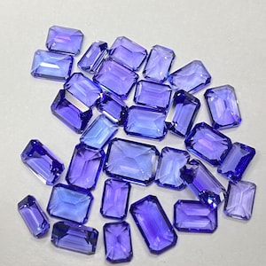 May include: A scattering of rectangular, cut gemstones in shades of blue and purple. The gems are of varying sizes and are displayed against a light background, highlighting their clarity and sparkle. These gems are likely for jewellery making.