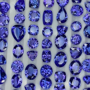 May include: A collection of polished, deep blue gemstones in various shapes, including oval, pear, and cushion cuts. The gems are arranged closely together, showcasing their facets and brilliance. The collection highlights the rich colour and clarity of the stones.