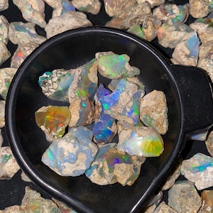 May include: A black bowl containing rough opal gemstones, displaying iridescent colours. The opals exhibit a spectrum of colours, including blue, green, and orange, set against a background of smaller, raw stones. The bowl is surrounded by more raw opal stones.