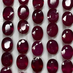 May include: A collection of deep red, oval-cut rubies arranged in five vertical rows on a white display tray. Each gemstone features a faceted surface that reflects light, showcasing a consistent crimson hue and a polished, symmetrical shape.