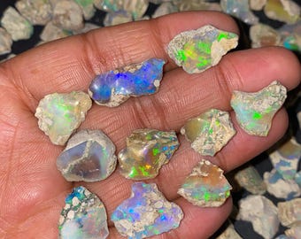 Puede incluir: Una colección de piedras preciosas de ópalo en bruto, que muestra una variedad de colores, incluyendo azules, verdes y naranjas. Los ópalos se sostienen en una mano, con muchos más dispersos en el fondo. Las piedras en bruto tienen un aspecto natural y sin pulir.