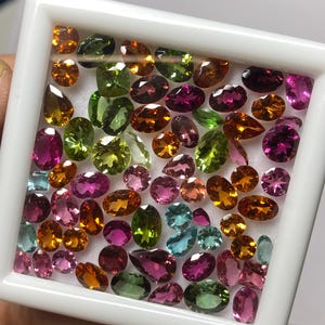 May include: A collection of colorful gemstones in a white tray. The gems are various shapes and sizes, including oval, pear, and round cuts. Colors include shades of orange, green, pink, purple, and blue. The gems are displayed in a white, square tray.