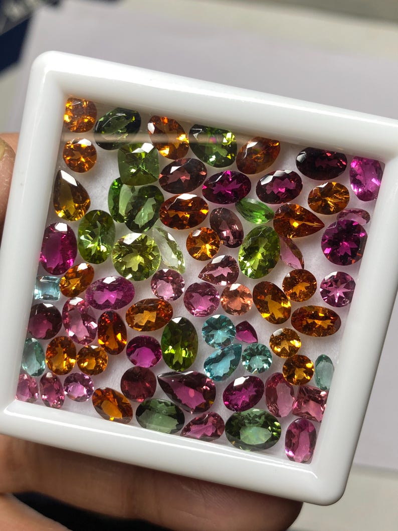 May include: A collection of colorful gemstones in a white tray. The gems are various shapes and sizes, including oval, pear, and round cuts. Colors include shades of orange, green, pink, purple, and blue. The gems are displayed in a white, square tray.