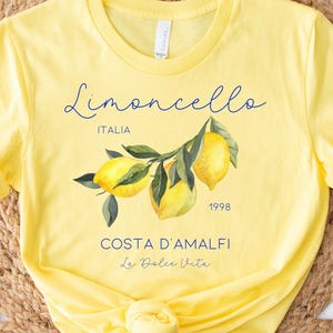 Puede incluir: Camiseta amarilla con una ilustraci&oacute;n de acuarela de limones en una rama. La camiseta presenta el texto "Limoncello" en cursiva, junto con "ITALIA", "1998", "COSTA D'AMALFI" y "La Dolce Vita".