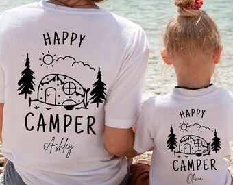 May include: White t-shirts with a black graphic of a camper, trees, sun, and clouds. The text "HAPPY CAMPER" is above the graphic, with a name below. The shirts are worn by two people sitting on a rocky beach.