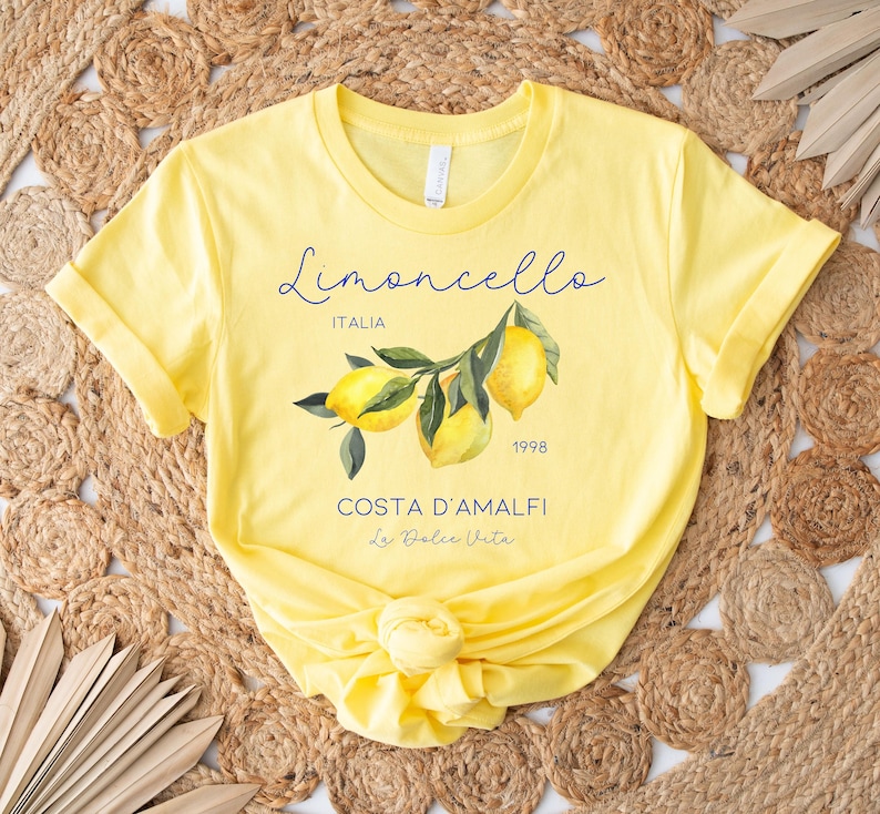 Puede incluir: Camiseta amarilla con una ilustraci&oacute;n de acuarela de limones en una rama. La camiseta presenta el texto "Limoncello" en cursiva, junto con "ITALIA", "1998", "COSTA D'AMALFI" y "La Dolce Vita".