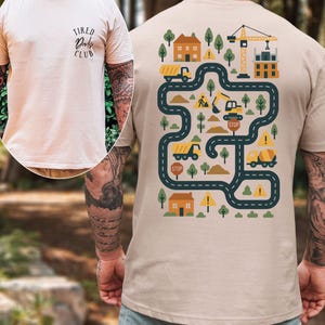 Może przedstawiać: T-shirt w kolorze piaskowym z grafiką o tematyce budowlanej na plecach, przedstawiającą drogę, ciężar&oacute;wki, domy, drzewa i dźwig. Prz&oacute;d koszulki zawiera napis "Tired Dads Club".