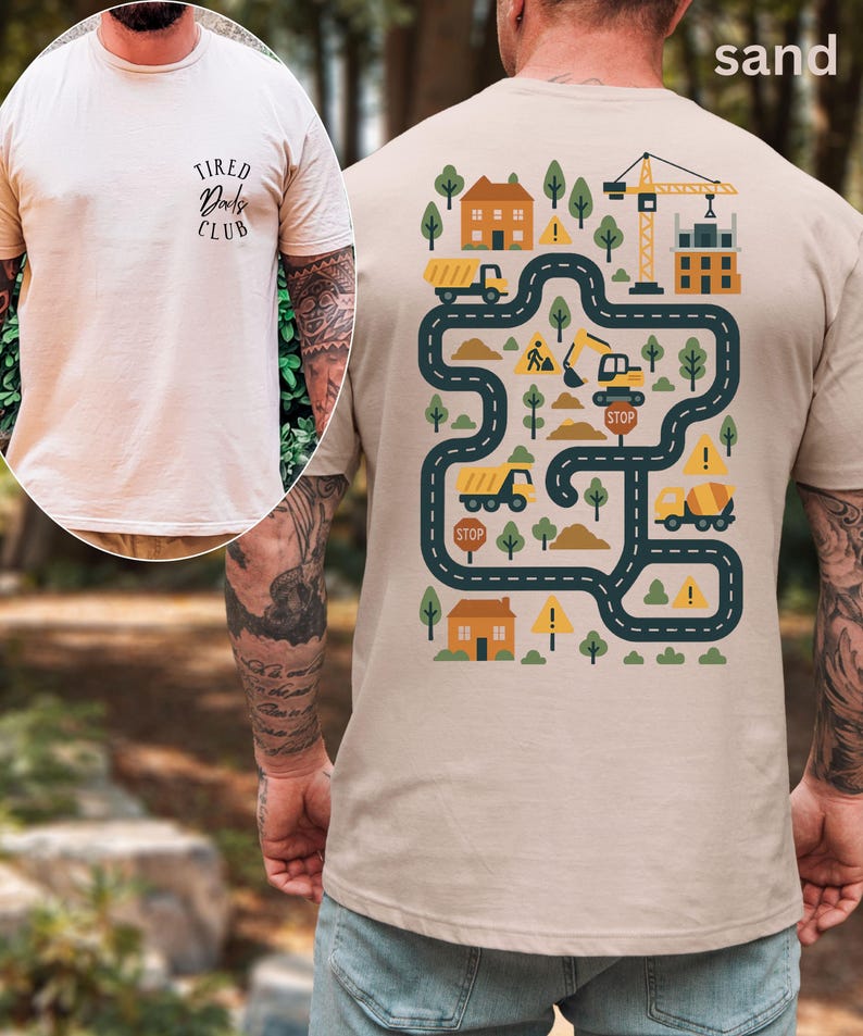 Może przedstawiać: T-shirt w kolorze piaskowym z grafiką o tematyce budowlanej na plecach, przedstawiającą drogę, ciężar&oacute;wki, domy, drzewa i dźwig. Prz&oacute;d koszulki zawiera napis "Tired Dads Club".