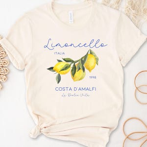 Puede incluir: Camiseta color crema con una ilustraci&oacute;n de acuarela de limones en una rama. La camiseta tiene la palabra "Limoncello" en cursiva, con "ITALIA" debajo. Texto adicional incluye "COSTA D'AMALFI" y "La Dolce Vita".