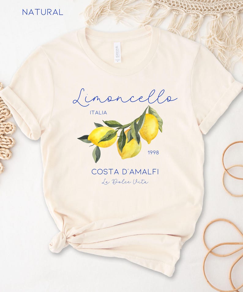 Puede incluir: Camiseta color crema con una ilustraci&oacute;n de acuarela de limones en una rama. La camiseta tiene la palabra "Limoncello" en cursiva, con "ITALIA" debajo. Texto adicional incluye "COSTA D'AMALFI" y "La Dolce Vita".
