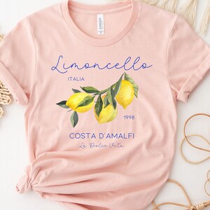 Puede incluir: Camiseta melocot&oacute;n con una ilustraci&oacute;n de limones en una rama. La camiseta presenta el texto "Limoncello ITALIA 1998 COSTA D'AMALFI La Dolce Vita" en escritura azul. La camiseta est&aacute; hecha de material suave. Arriba est&aacute; "Heather Peach".