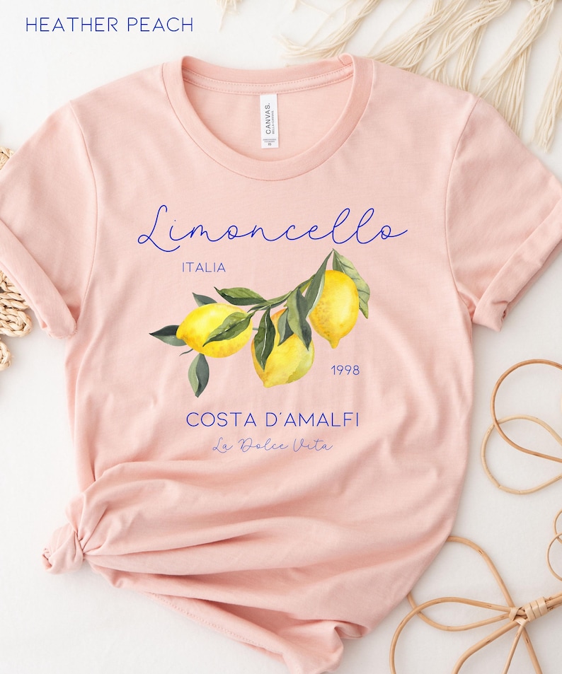 Puede incluir: Camiseta melocot&oacute;n con una ilustraci&oacute;n de limones en una rama. La camiseta presenta el texto "Limoncello ITALIA 1998 COSTA D'AMALFI La Dolce Vita" en escritura azul. La camiseta est&aacute; hecha de material suave. Arriba est&aacute; "Heather Peach".