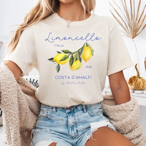 Puede incluir: Camiseta color crema con el texto "Limoncello Italia 1998 Costa D'Amalfi La Dolce Vita" y una ilustraci&oacute;n de limones. La camiseta se combina con pantalones cortos vaqueros.