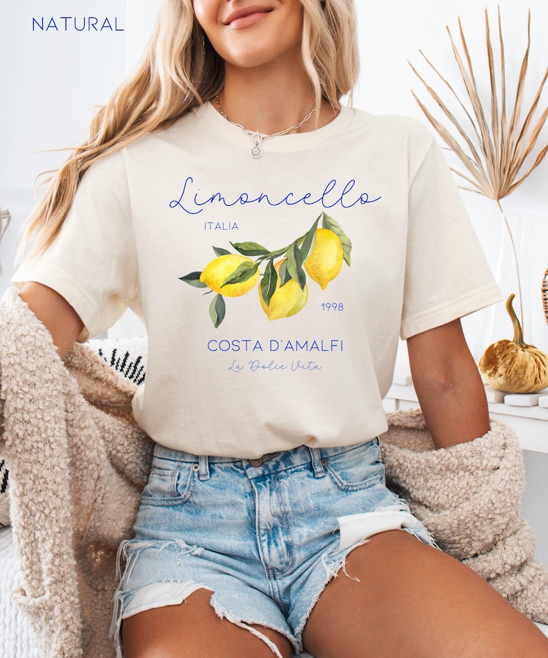 Puede incluir: Camiseta color crema con el texto "Limoncello Italia 1998 Costa D'Amalfi La Dolce Vita" y una ilustraci&oacute;n de limones. La camiseta se combina con pantalones cortos vaqueros.