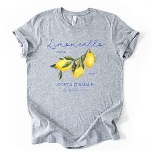 Puede incluir: Camiseta gris con la frase "Limoncello Italia 1998 Costa D'Amalfi" en azul. La camiseta presenta una ilustraci&oacute;n de acuarela de limones en una rama. El texto "Athletic Heather" est&aacute; en la parte superior.