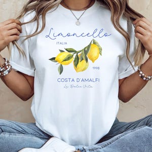 Puede incluir: Camiseta blanca con una ilustraci&oacute;n de limones y el texto "Limoncello Italia 1998" en azul. Debajo, se lee "Costa D'Amalfi La Dolce Vita". La palabra "WHITE" est&aacute; en la esquina superior izquierda.