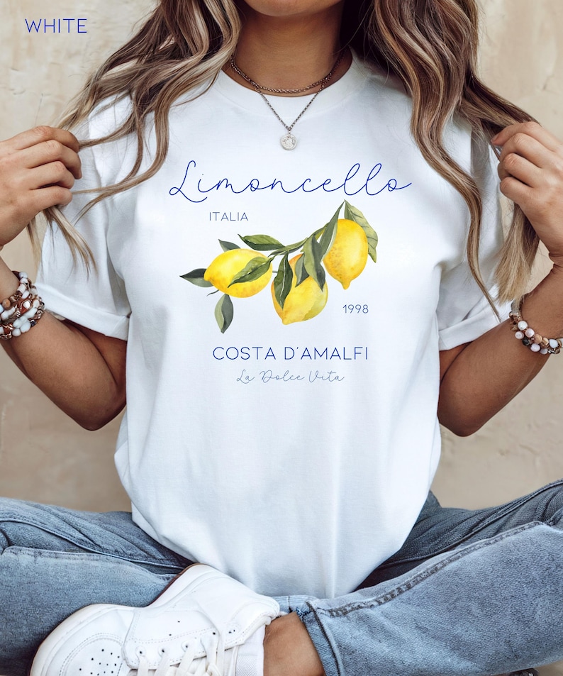 Puede incluir: Camiseta blanca con una ilustraci&oacute;n de limones y el texto "Limoncello Italia 1998" en azul. Debajo, se lee "Costa D'Amalfi La Dolce Vita". La palabra "WHITE" est&aacute; en la esquina superior izquierda.