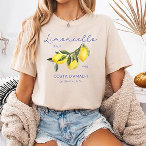 Puede incluir: Camiseta de color crema suave con una ilustraci&oacute;n de acuarela de limones en una rama. La camiseta tiene el texto "Limoncello ITALIA 1998 COSTA D'AMALFI La Dolce Vita" en escritura azul. La camiseta es de manga corta.