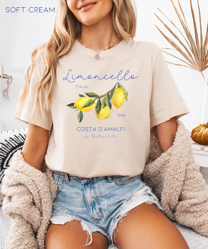 Puede incluir: Camiseta de color crema suave con una ilustraci&oacute;n de acuarela de limones en una rama. La camiseta tiene el texto "Limoncello ITALIA 1998 COSTA D'AMALFI La Dolce Vita" en escritura azul. La camiseta es de manga corta.