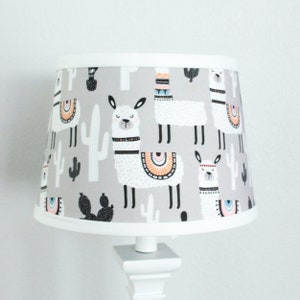 May include: A gray lampshade with a white border features a repeating pattern of white llamas with black accents and colorful details, cacti, and a gray background.