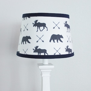 May include: A white lampshade with a navy blue trim features a repeating pattern of moose, bears, and crossed arrows. The lampshade is on a white lamp base.