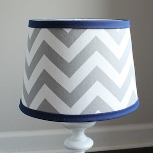 May include: A white lampshade with a gray and white chevron pattern and a navy blue trim. The lampshade is on a white lamp base.