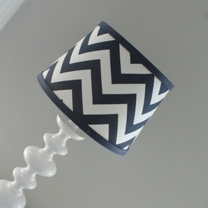K&ouml;nnte beinhalten: Wei&szlig;e Lampe mit marineblau-wei&szlig;em Chevron-Lampenschirm. Die Lampe hat einen wei&szlig;en, geschwungenen Sockel. Der Lampenschirm ist mit marineblauem Stoff eingefasst. Dekorativer Beleuchtungsartikel.