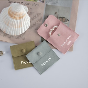 May include: A selection of small, suede-like pouches in pink, green, and grey, each with a snap closure. One pink pouch contains a pearl bracelet. The pouches are labelled with names in white script: "Maid of honor Gilda", "Dove", and "Jannah".