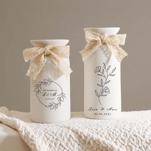 May include: Two white ceramic vases with cream-colored lace bows at the top. One vase has a floral wreath design with the text "THE WEDDING OF L&M 03.06.2025". The other vase has a floral branch design with the text "Lara & Marc 03.06.2025".
