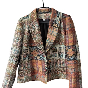 Könnte beinhalten: Ein gemusterter Blazer mit Patchwork-Design in Braun-, Grün- und Orangetönen. Der Blazer hat ein schwarzes Futter und einen einzelnen Knopfverschluss. Der Stoff weist Blumen- und geometrische Muster auf.