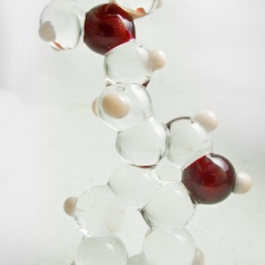 May include: A glass model of a molecule with clear, white, and red beads. The beads are connected by clear rods. The model is held by a finger.
