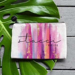 May include: A white canvas zippered pouch with a pink, purple, and gold abstract watercolor design. The text "stacci BRIDE" is printed on the pouch.